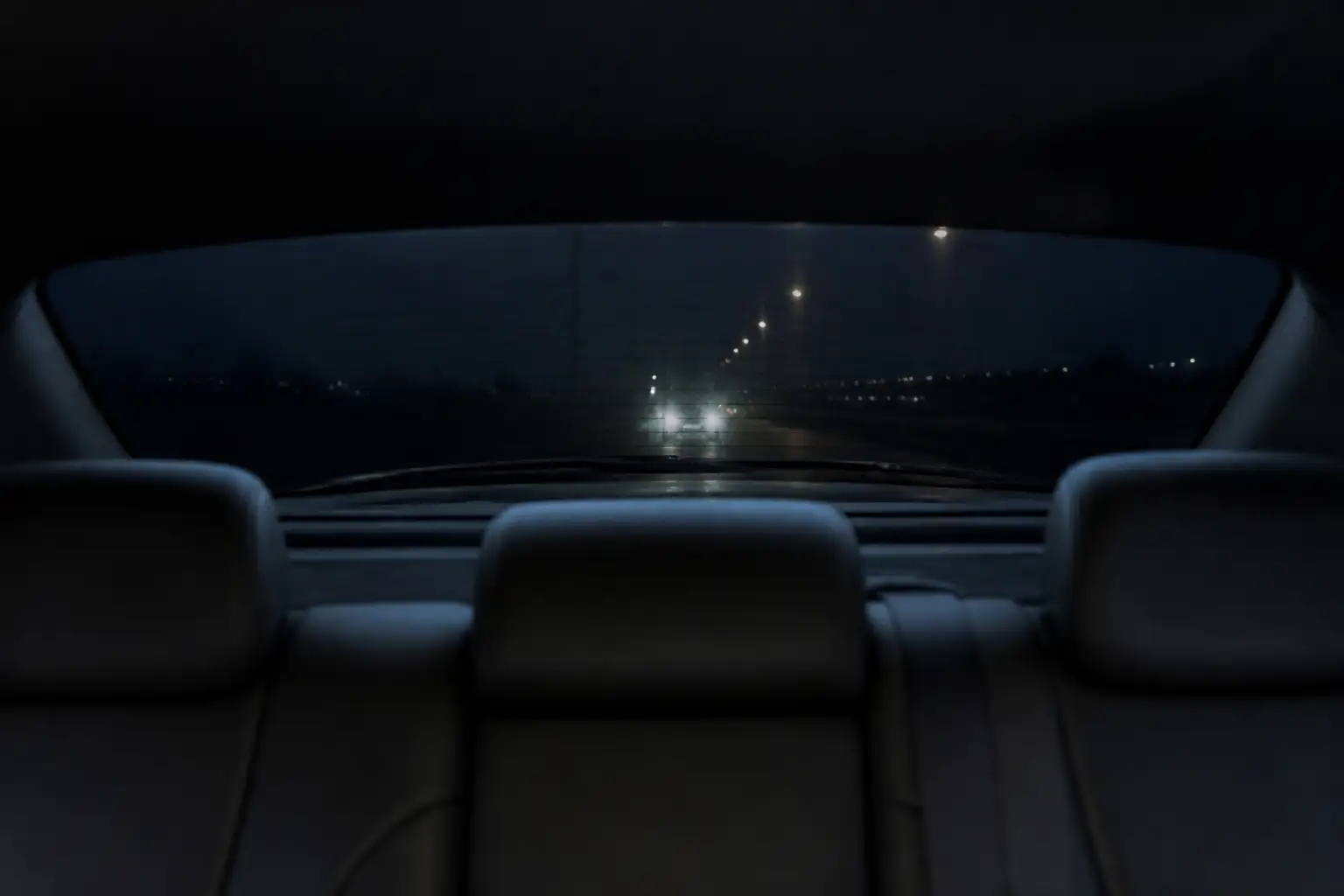 Blick von der Rückbank durch die getönte Heckscheibe bei Nacht, die Scheinwerfer eines folgenden Fahrzeugs und Straßenlaternen erscheinen nur noch als Lichtpunkte
