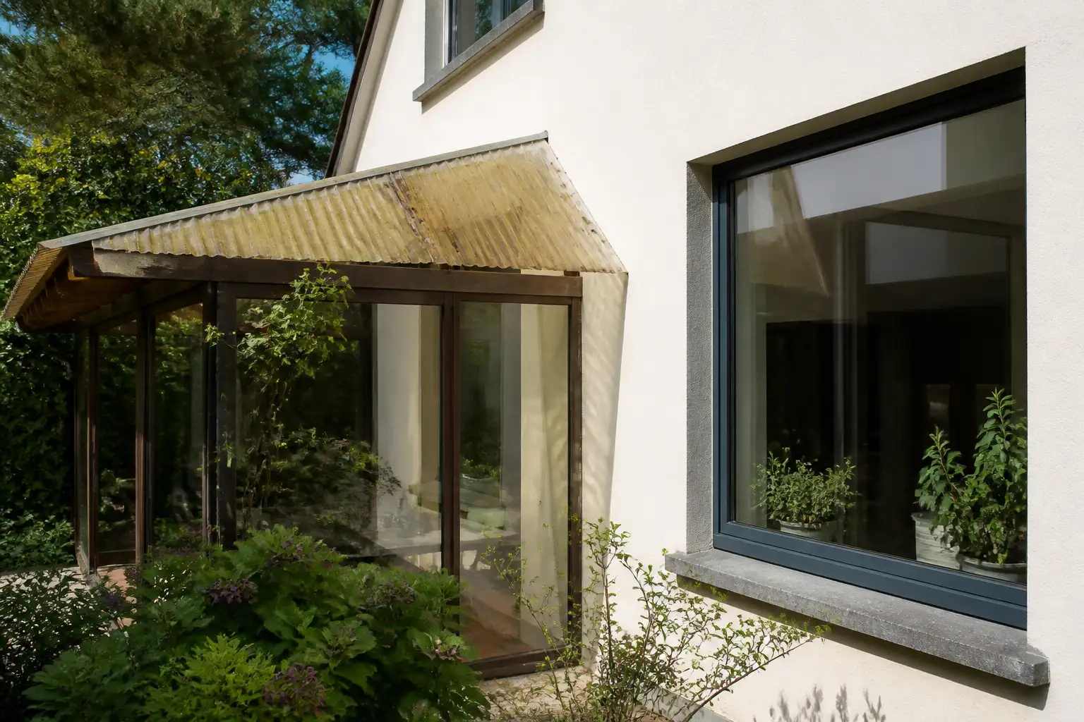 Hausecke mit Terrassenvordach aus gewelltem Kunststoff, das gelblich und trüb geworden ist, daneben ein großes Fenster mit klarem Glas